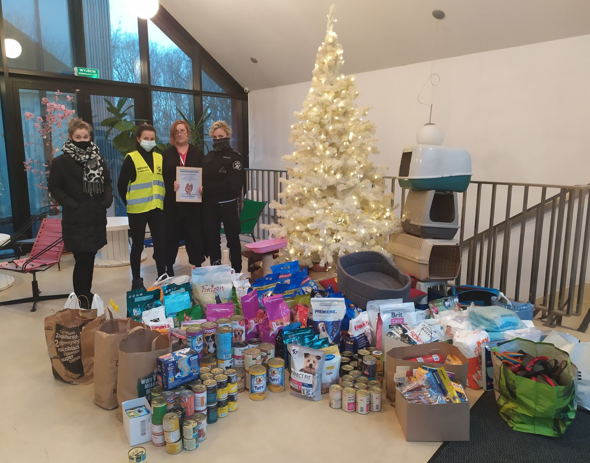 Zdjęcie dwóch pracowników SDK wraz z dwoma pracownikami OTOZ Animals. a pierwszym planie wszystkie dary ze zbiórki. Koordynatorka trzyma pamiątkowe podziękowania. W tle biała oświetlona choinka