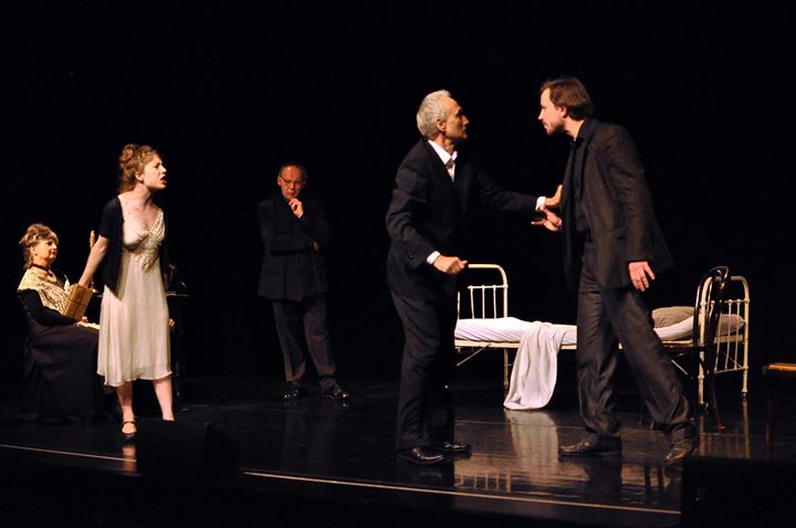 Zdjęcie promocyjne pracowni.  Zdjęcie wykonane w trakcie przedstawienia. Na zdjęciu aktorzy na scenie teatralnej. Mężczyzna na pierwszym rozmawia z innym mężczyzną. Obok stoi kobieta. W tle pozostali aktorzy.