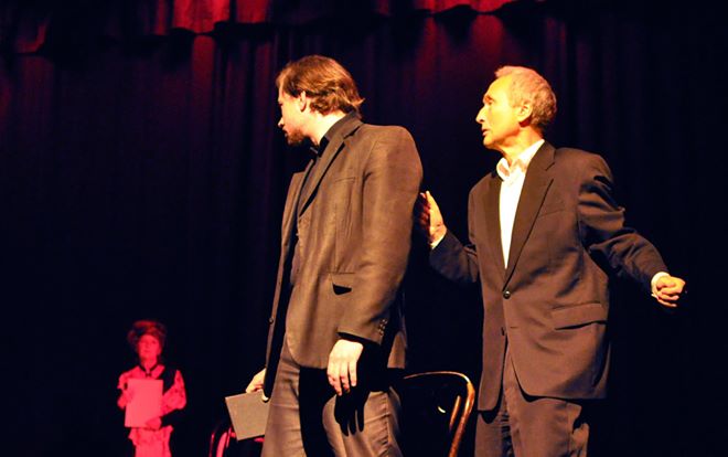 Zdjęcie promocyjne pracowni. Zdjęcie wykonane w trakcie przedstawienia. Na zdjęciu aktorzy na scenie teatralnej. Dwóch mężczyzn na pierwszym planie rozmawia ze sobą.