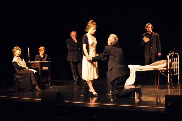 Zdjęcie promocyjne pracowni. Zdjęcie wykonane w trakcie przedstawienia. Na zdjęciu aktorzy na scenie teatralnej. Mężczyzna na pierwszym planie klęczy przed kobietą. W tle pozostali aktorzy.