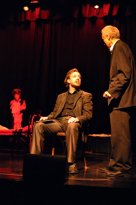 Zdjęcie promocyjne pracowni.  Zdjęcie wykonane w trakcie przedstawienia. Na zdjęciu siedzący aktor na fotelu. Obok niego stoi drugi mężczyzna.