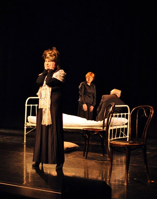 Zdjęcie promocyjne pracowni.  Zdjęcie wykonane w trakcie przedstawienia. Na zdjęciu stojąca kobieta z rękami złożonymi przy twarzy. W tle, na łóżku siedzi mężczyzna, obok niego stoi druga kobieta.