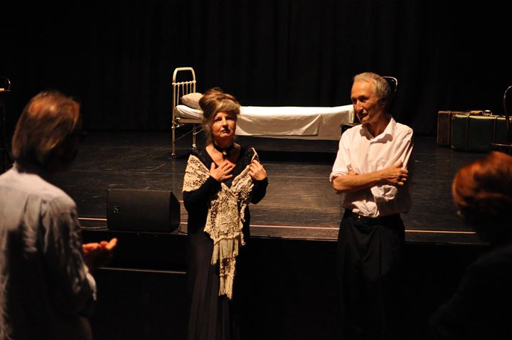 Zdjęcie promocyjne pracowni.  Zdjęcie wykonane w trakcie przedstawienia. Na zdjęciu kobieta oraz mężczyzna stoją przed sceną. W tle widoczne łóżko stojące na scenie.